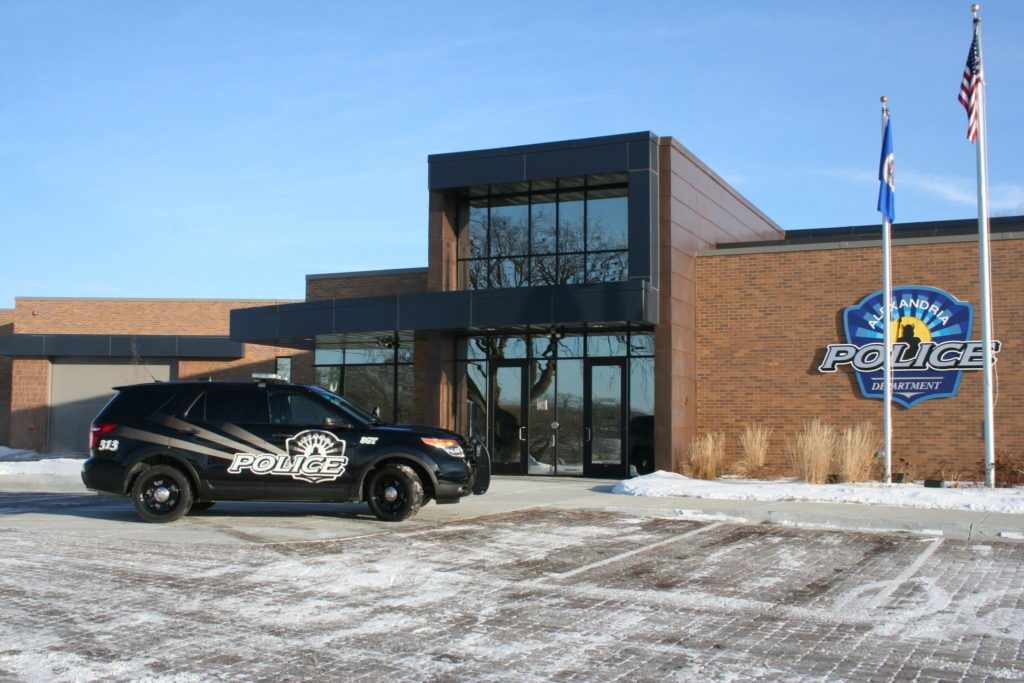 A black police SUV is parked in front of the modern brick and glass Alexandria Police Department building. Light snow dusts the area, emphasizing the dedication to public safety. Two flags on poles proudly stand near the entrance, symbolizing law enforcement's watchful presence.