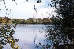 A tall blue water tower stands in the distance, reflected in a calm lake within the serene city park. Trees frame the scene, with clear skies above and a row of small birds gliding on the water, creating a picturesque backdrop for nearby trails.