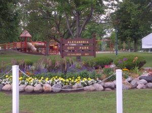 A park scene with a sign reading "Alexandria City Park, Established 1921." In the background, there is a children's play area with playground equipment. The foreground features colorful flowers and a white rope fence, part of the scenic trails that make city parks beloved by all.