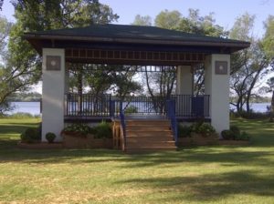 A small outdoor gazebo with a roof and open sides stands in a grassy city park. It has a staircase leading up and is surrounded by trees and trails. A body of water is visible in the background under a clear blue sky.