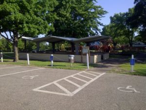 A city park with a covered pavilion, surrounded by trees and connecting trails. A playground with slides is visible in the background. In the foreground, a parking lot features accessible spaces. The scene is highlighted beautifully by daylight.