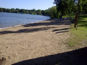 A sandy beach stretches along a serene lake with clear blue water, part of the city's beautiful parks and trails. Trees and greenery line the shore, casting gentle shadows on the sand. The sky is sunny, suggesting a pleasant day. Nearby, a bench and a few signs are nestled among the trees.