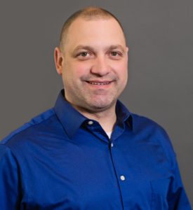 Andy Mellgren is smiling with short hair and wearing a blue button-up shirt. The background is a plain gray color.