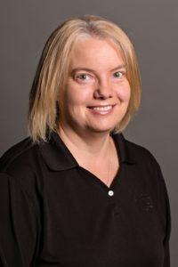 A woman with shoulder-length blonde hair smiles at the camera. She is wearing a black collared shirt and stands against a plain gray background.