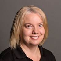 A woman with shoulder-length blonde hair smiles at the camera. She is wearing a black collared shirt and stands against a plain gray background.