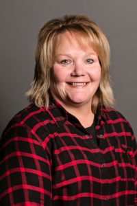 Smiling woman with shoulder-length blond hair, wearing a black and red plaid shirt, posed in front of a gray background.