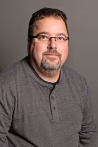 A man, resembling Mike Schmidt, with short dark hair and glasses wears a gray long-sleeve shirt and sports a beard. Seated against a plain gray background, he looks directly at the camera with a neutral expression.