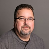 A man, resembling Mike Schmidt, with short dark hair and glasses wears a gray long-sleeve shirt and sports a beard. Seated against a plain gray background, he looks directly at the camera with a neutral expression.