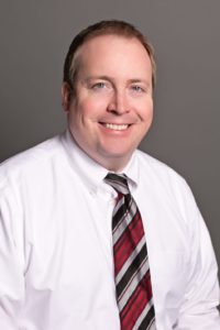 Martin Schultz, a smiling man in a white shirt and striped tie, poses confidently against a gray background.