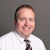 Martin Schultz, a smiling man in a white shirt and striped tie, poses confidently against a gray background.