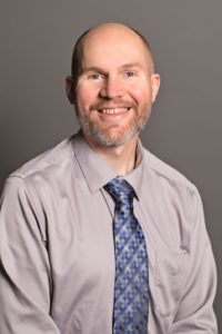 A smiling Reed Heidelberger, with a bald head and beard, is elegantly dressed in a light gray shirt paired with a blue patterned tie, posing against a plain gray background.