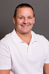 Bill Thoennes is smiling warmly while wearing a white polo shirt against a gray background.