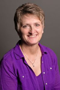 Smiling woman with short hair wearing a purple shirt and necklace, seated against a gray background.