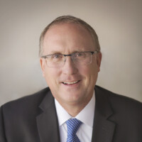 A man in a black suit and blue tie smiles warmly at the camera, his glasses perched neatly on his nose. With short, light hair, Thomas Jacobson stands against a neutral, blurred background.