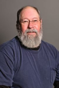 A man with a gray beard and glasses, possibly Vinnie Hennen, is wearing a navy blue shirt. He is looking at the camera, smiling slightly against a plain gray background.