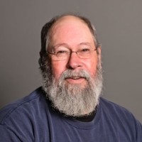 A man with a gray beard and glasses, possibly Vinnie Hennen, is wearing a navy blue shirt. He is looking at the camera, smiling slightly against a plain gray background.