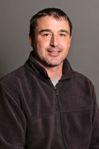 A person with short dark hair and light facial hair is smiling. They are wearing a dark zip-up fleece over a gray shirt. The background is a plain gray.