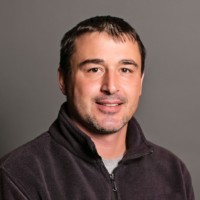 A person with short dark hair and light facial hair is smiling. They are wearing a dark zip-up fleece over a gray shirt. The background is a plain gray.