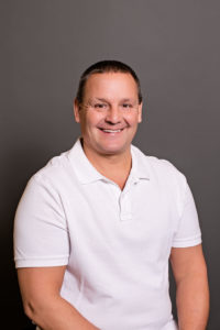 A person with short dark hair is smiling while sitting against a plain dark gray background. They are wearing a white polo shirt.