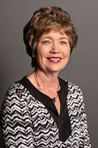 Erin Anderson, a smiling woman with short, curly hair, wears a black and white patterned top and a necklace, posed against a plain gray background.