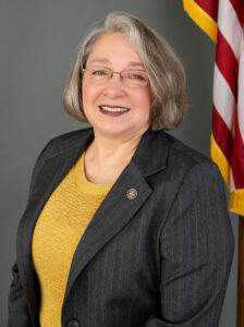 Bobbie Osterberg, a smiling figure with short gray hair and glasses, wears a dark blazer adorned with a pin and a mustard-colored top. They stand proudly in front of an American flag, set against a gray background.