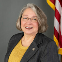Bobbie Osterberg, a smiling figure with short gray hair and glasses, wears a dark blazer adorned with a pin and a mustard-colored top. They stand proudly in front of an American flag, set against a gray background.