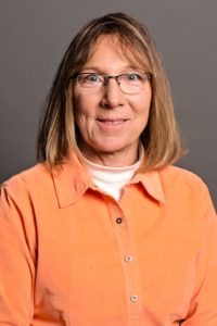 A woman with shoulder-length brown hair and glasses is wearing an orange button-up shirt over a white turtleneck. She is smiling slightly, and the background is a plain gray.