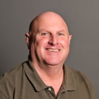 A smiling man with a bald head is wearing a green collared shirt. He is posed against a plain gray background.