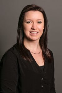 A woman with long, dark brown hair wearing a black shirt and necklace smiles at the camera against a plain gray background.