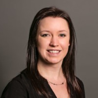 A woman with long, dark brown hair wearing a black shirt and necklace smiles at the camera against a plain gray background.