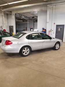 A silver sedan is parked inside a spacious garage with concrete flooring and white walls, hinting at a space prepped for competitive bidding. The car faces away from the camera, with equipment surrounding it and a partially open garage door in the background.