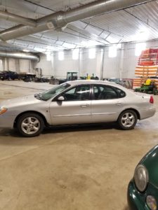 A silver sedan is parked indoors on a concrete floor, surrounded by other vehicles, a forklift, and stacked red and white barriers. This large garage or warehouse, ideal for auction events, often features visible ductwork as bidders familiarize themselves with the auction terms.