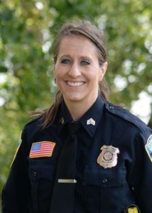 In this Employee Spotlight, Sgt. Tina Lake is seen outdoors, proudly wearing her police uniform adorned with badges and an American flag patch. With her long hair pulled back and a smile on her face, she stands against a backdrop of blurred trees.