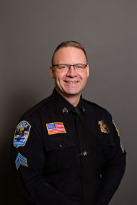 Sergeant Kevin Guenther, featured in our Employee Spotlight, stands smiling against a gray background. Dressed in a police uniform adorned with patches like the American flag and badge, he exudes professionalism. His glasses and short hair add to his distinguished appearance.