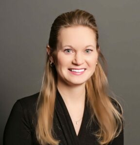 Karin Tank, featured in our Employee Spotlight, smiles while flaunting her long, light brown hair in a chic black top against a gray backdrop.