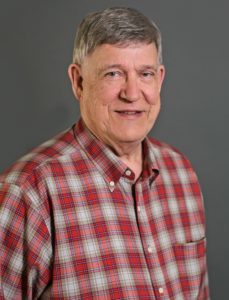 An older adult with short gray hair, Roger Thalman, is smiling at the camera. He wears a red and gray plaid button-up shirt against a plain gray background.