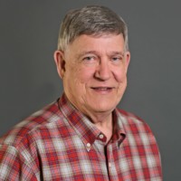An older adult with short gray hair, Roger Thalman, is smiling at the camera. He wears a red and gray plaid button-up shirt against a plain gray background.