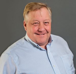 Smiling man with short, light hair in a white and blue checkered shirt against a gray background, reminiscent of Bill Franzen's relaxed style.