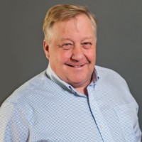 Smiling man with short, light hair in a white and blue checkered shirt against a gray background, reminiscent of Bill Franzen's relaxed style.
