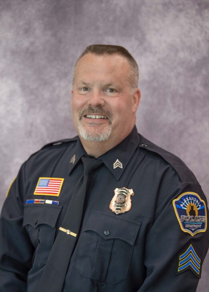 Police officer Chad Schroeder, in a dark blue uniform adorned with badges and patches, smiles warmly in front of a mottled gray and purple backdrop.