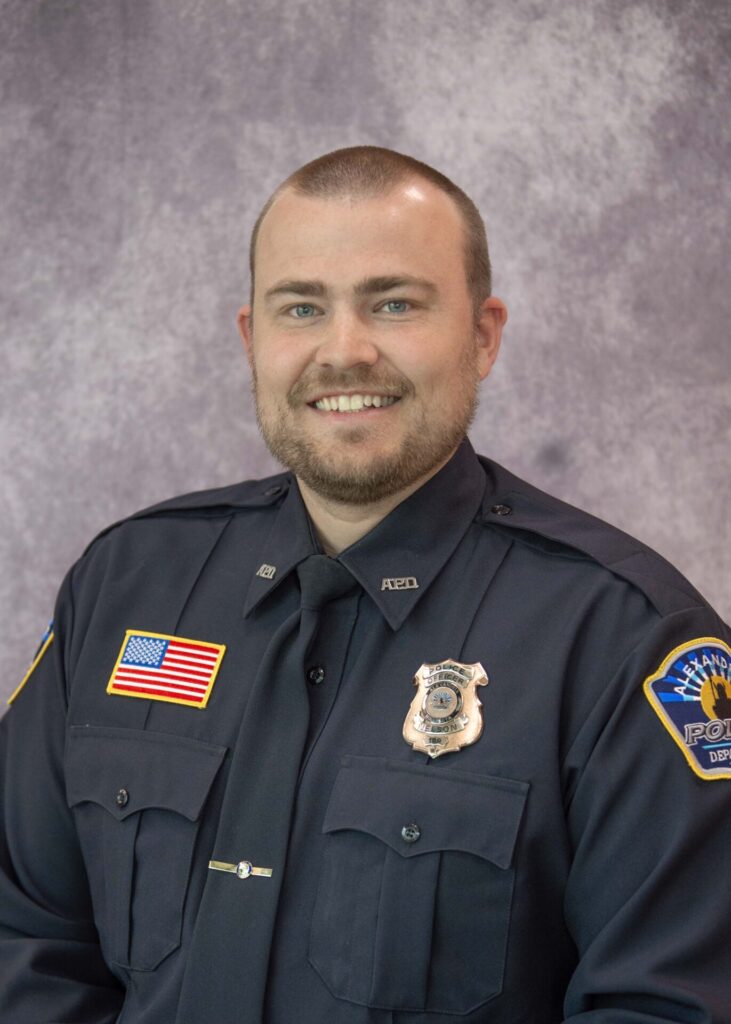 A smiling police officer in a dark uniform poses in front of a mottled gray background. His uniform features a U.S. flag patch, badge, and police department shoulder patch.