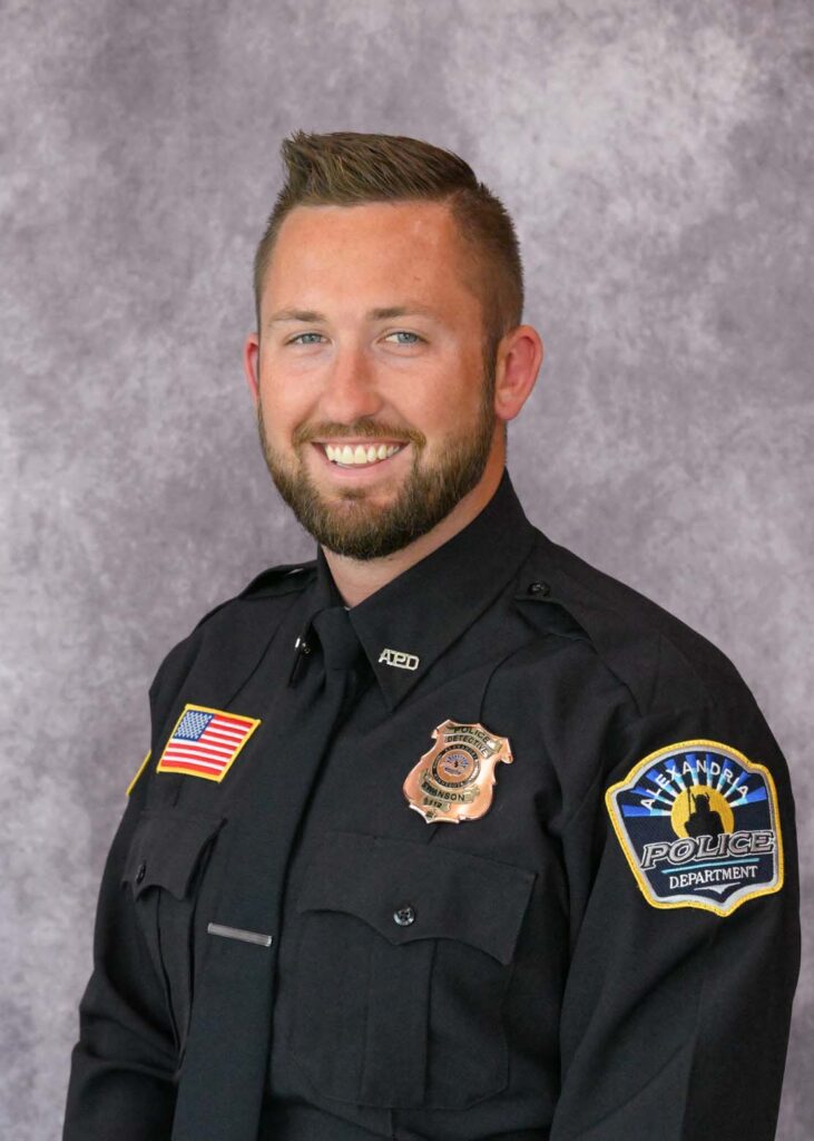 A smiling male police officer, identified as Alexander Swanson, with short brown hair and a beard wears a black uniform with badges and an American flag patch, posing in front of a light gray mottled background.