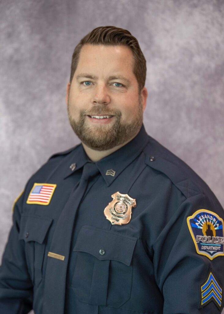 A male police officer, Brian Kakach, in a dark blue uniform with badges and an American flag patch, poses and smiles in front of a mottled gray background.