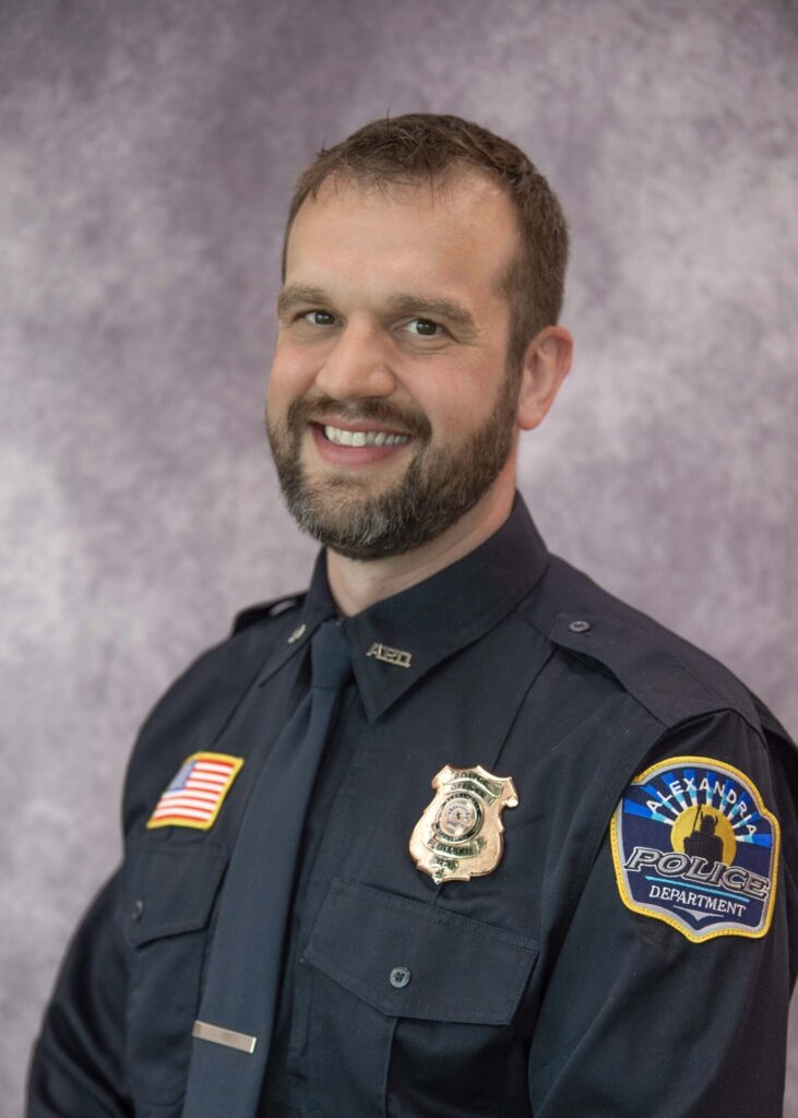 A smiling police officer with a beard, Brandon Plumski, poses for a portrait in uniform against a mottled gray background. His shirt has an American flag patch and a badge reading "Alexandria Police Department.