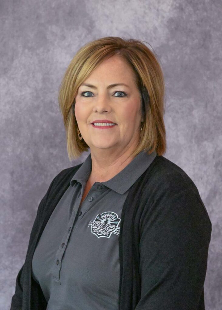 Cheryl Meyers, a woman with short blonde hair, wears a gray collared shirt featuring a police department logo and a black cardigan, posing confidently in front of a gray textured background.