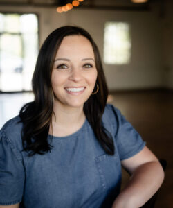 Kelsey Deling, a woman with long dark hair and hoop earrings, smiles at the camera. She wears a short-sleeved denim top against a softly lit background with warm tones and blurred elements, including a window.