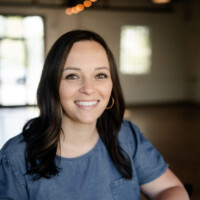 Kelsey Deling, a woman with long dark hair and hoop earrings, smiles at the camera. She wears a short-sleeved denim top against a softly lit background with warm tones and blurred elements, including a window.