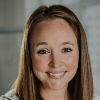 Heather Koskiniemi, with straight light brown hair and a striped white shirt, smiles at the camera against a softly blurred indoor background.