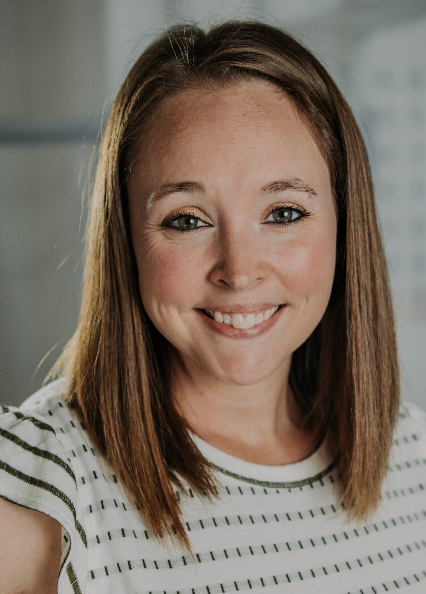 Heather Koskiniemi, with straight light brown hair and a striped white shirt, smiles at the camera against a softly blurred indoor background.