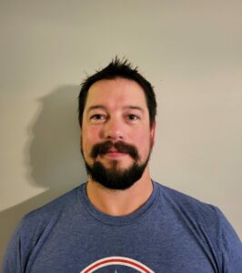 A man with short dark hair, a beard, and a mustache is wearing a blue T-shirt. He stands against a plain light-colored wall, smiling slightly at the camera.